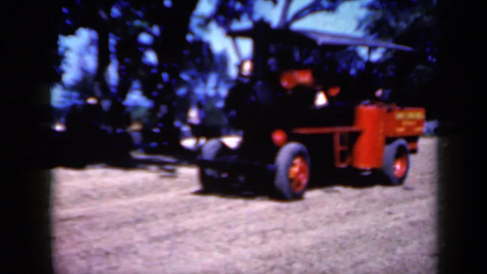 1959 Iowa footage frame with a black steam engine and red wheel assembly crossing a sunlit dirt field beneath trees.