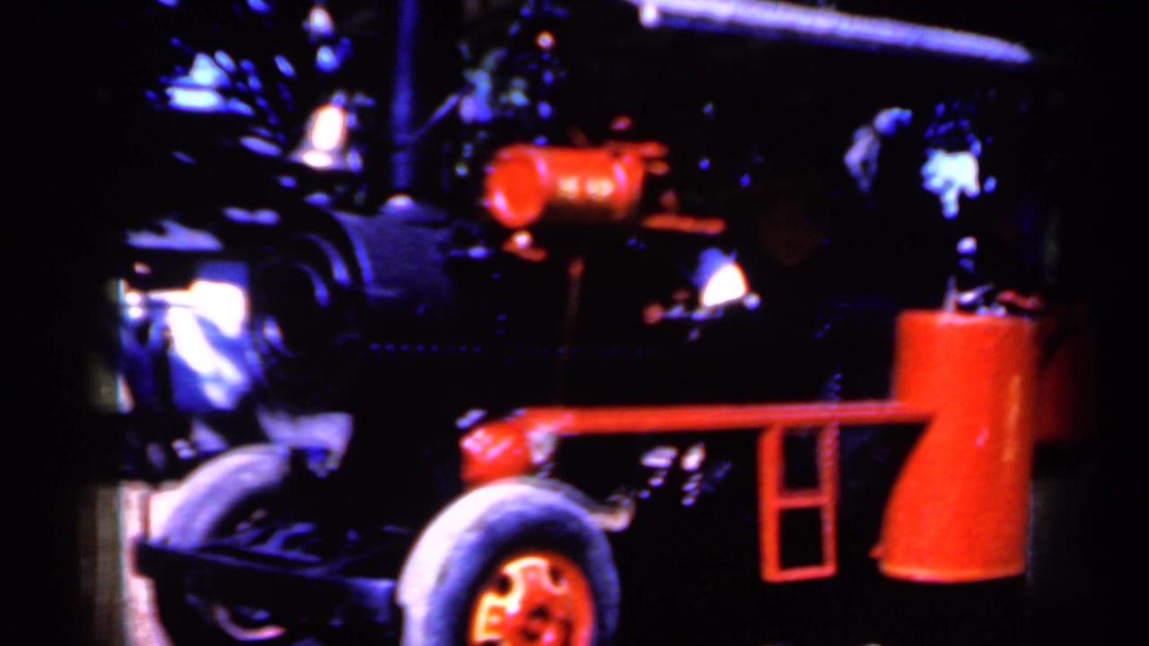 Small black steam engine with red-painted details beside leafy shade.