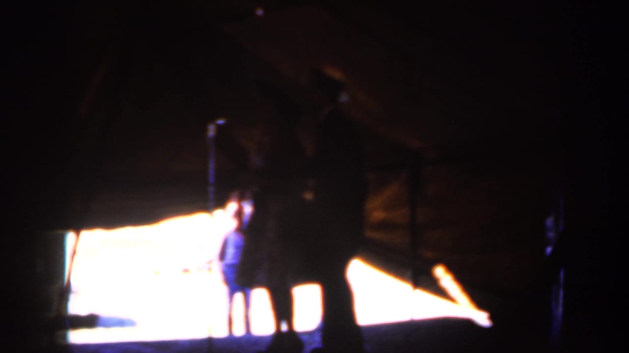 Dim 8mm frame with silhouetted visitors standing in front of a bright projected area, likely inside a railroad-station or visitor space.