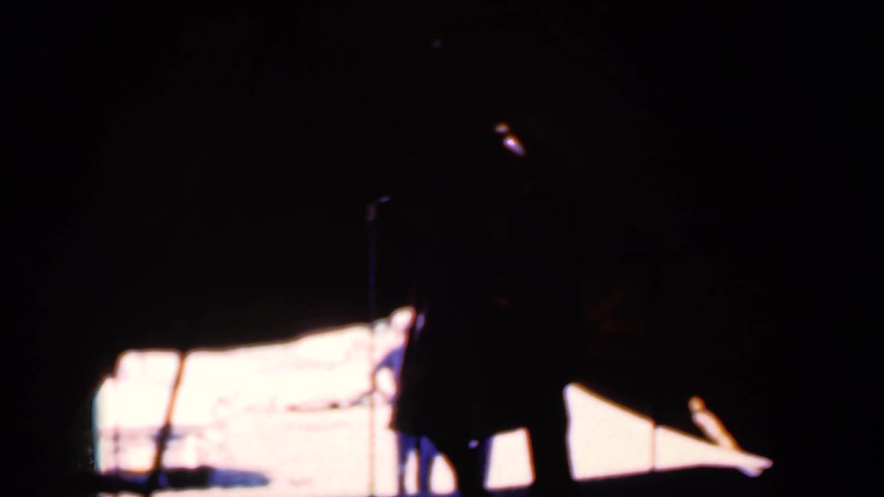 Dark dancer silhouettes stand against a bright stage backdrop under a tent.