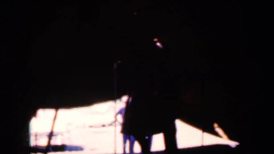 The dancers remain mostly in shadow, with the white tent-lit background and nearby microphone stand defining the stage area.