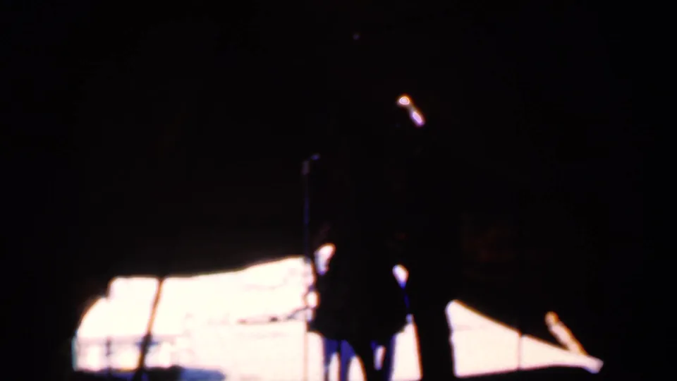 Two silhouetted performers are seen from the audience side, partly obscured by heavy shadow and bright outdoor light beyond the stage.