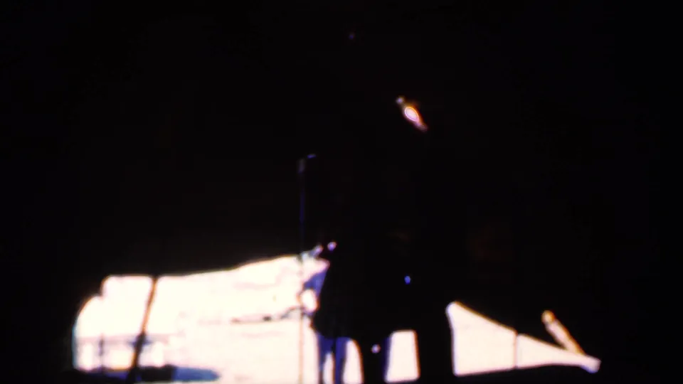 The final frame holds the couple in deep shadow beside a microphone stand, with bright sunlit tent fabric or opening behind them.