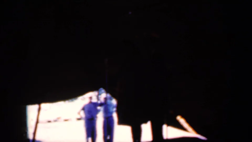In 1959 Iowa home-movie footage, two adults in blue clothing stand near a bright patch of light under a dark tent or canopy.