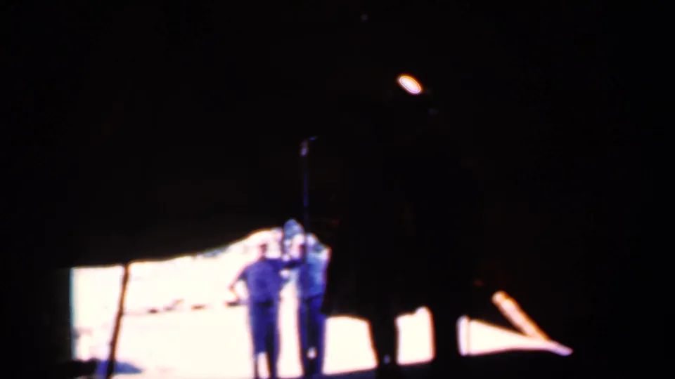 Two adults appear near a bright opening at night in the 1959 Iowa 8mm scene, framed by dark tent or canopy edges.