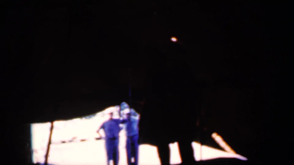 Two adults in blue clothing stand near a bright lit patch under a dark canopy in this 1959 Iowa home-movie frame.