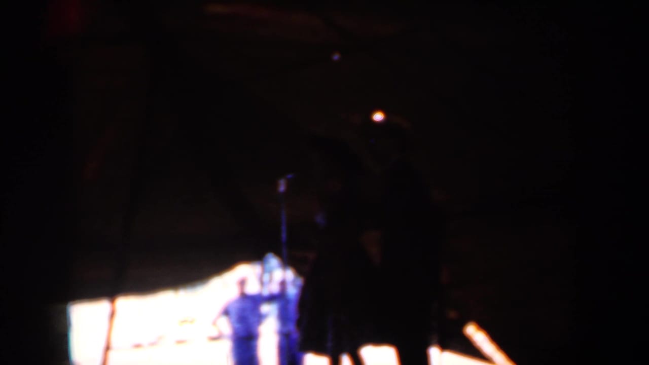 Dark tent scene with silhouetted figures near a brightly lit stage area.