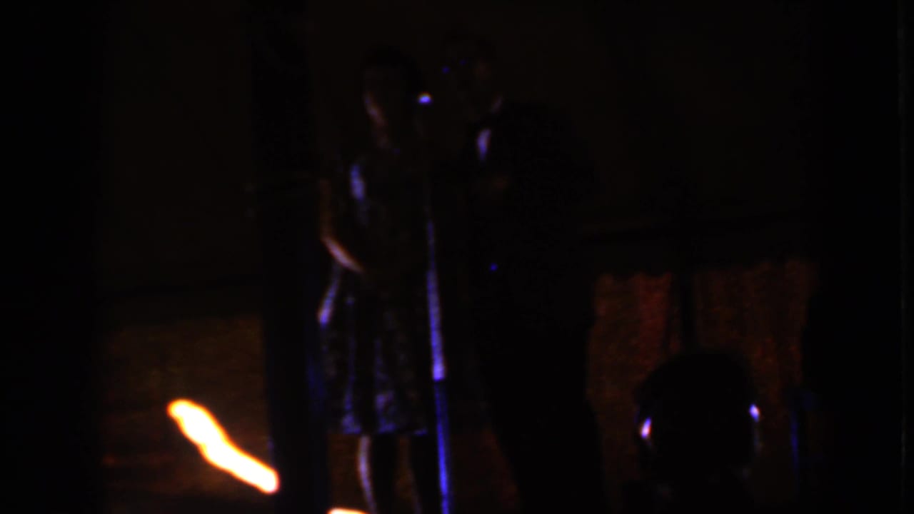 Dim stage with a dressed couple standing near a microphone and warm light at left.
