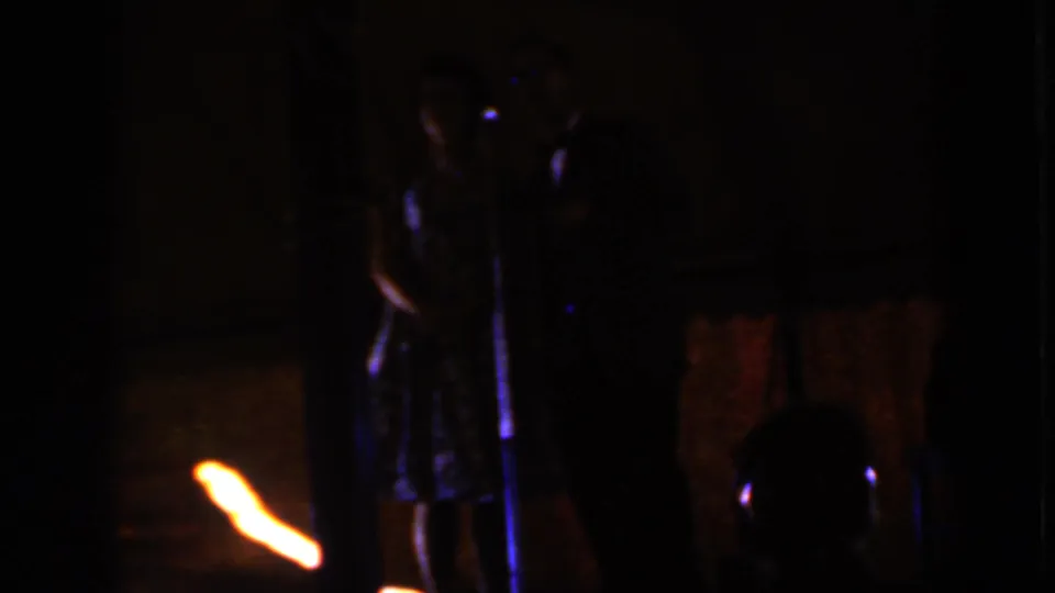 1959 Iowa 8mm frame showing a formally dressed couple on a dark stage near a standing microphone.