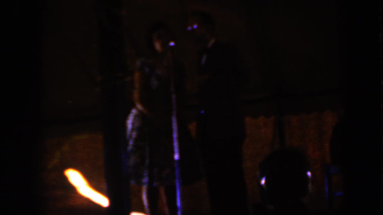 A formally dressed couple appears to address a room from a microphone on a low-lit Iowa stage in 1959.