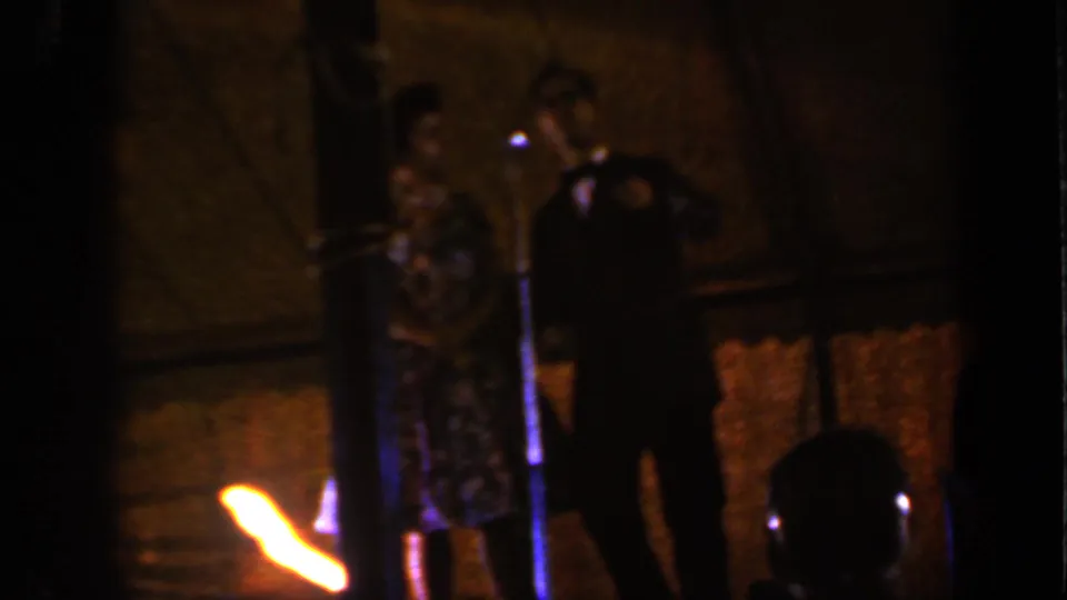 1959 Iowa home-movie frame of a woman in a patterned dress and a suited man standing at a stage microphone.