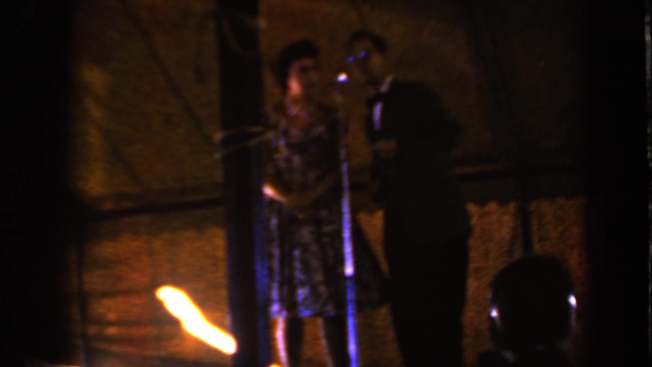 1959 Iowa home-movie frame showing two performers on a dark stage, one near a microphone, with a bright flame at lower left.