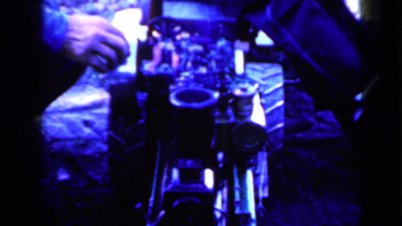 Blue-lit close view of a mechanical-looking object with hands and seated figures nearby.
