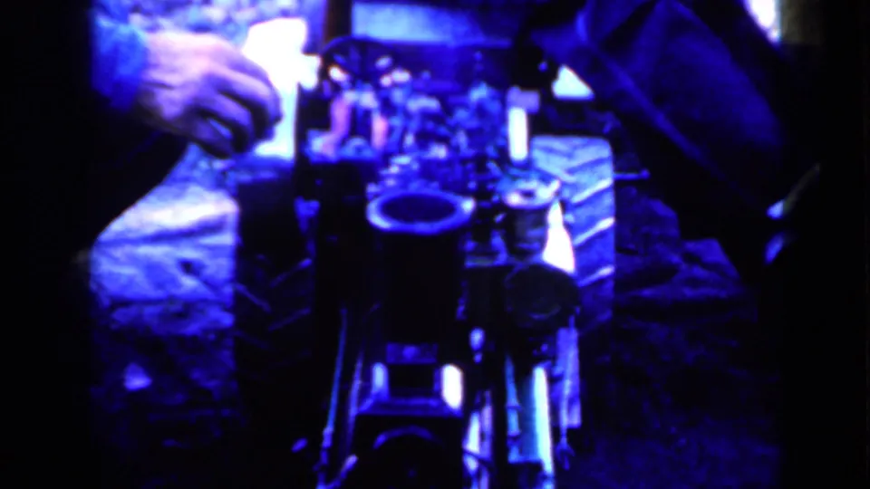 Blue-toned 1959 Iowa home-movie frame with hands near a mechanical-looking object or equipment, surrounded by seated figures.