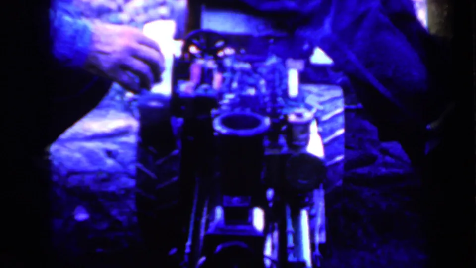 1959 Iowa home-movie frame showing a blue-lit close-up of hands beside equipment or a prop, with dark clothing at the edges.