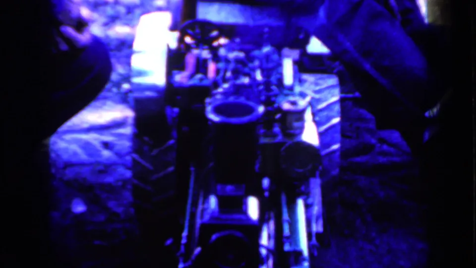 Blue-toned 1959 Iowa home-movie frame showing equipment or a stage prop from close range, with hands and seated people partly visible.