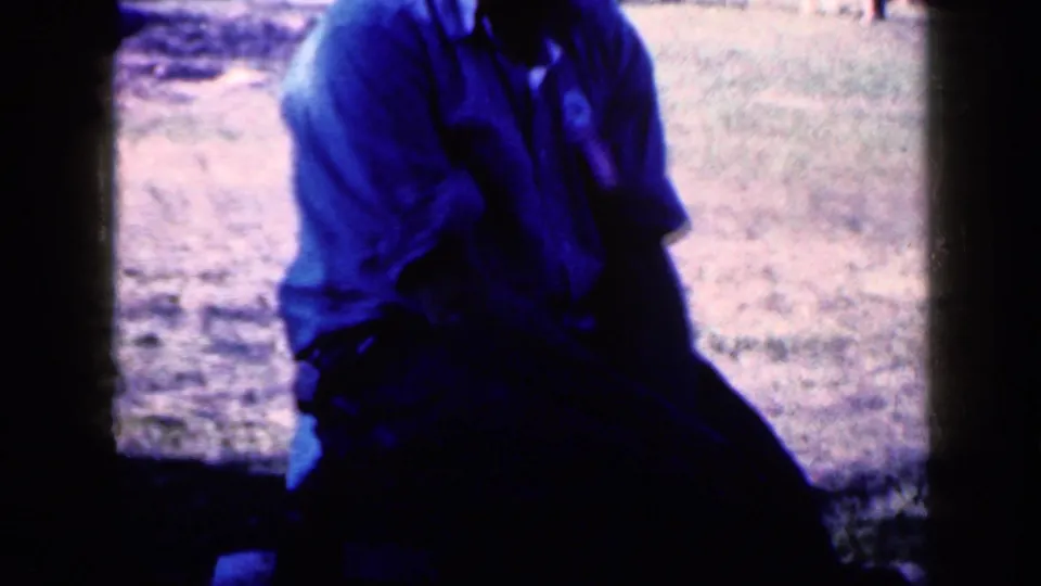 1959 Iowa 8mm frame showing an adult in a blue shirt seated outdoors on grass, with the face obscured by shadow and crop.