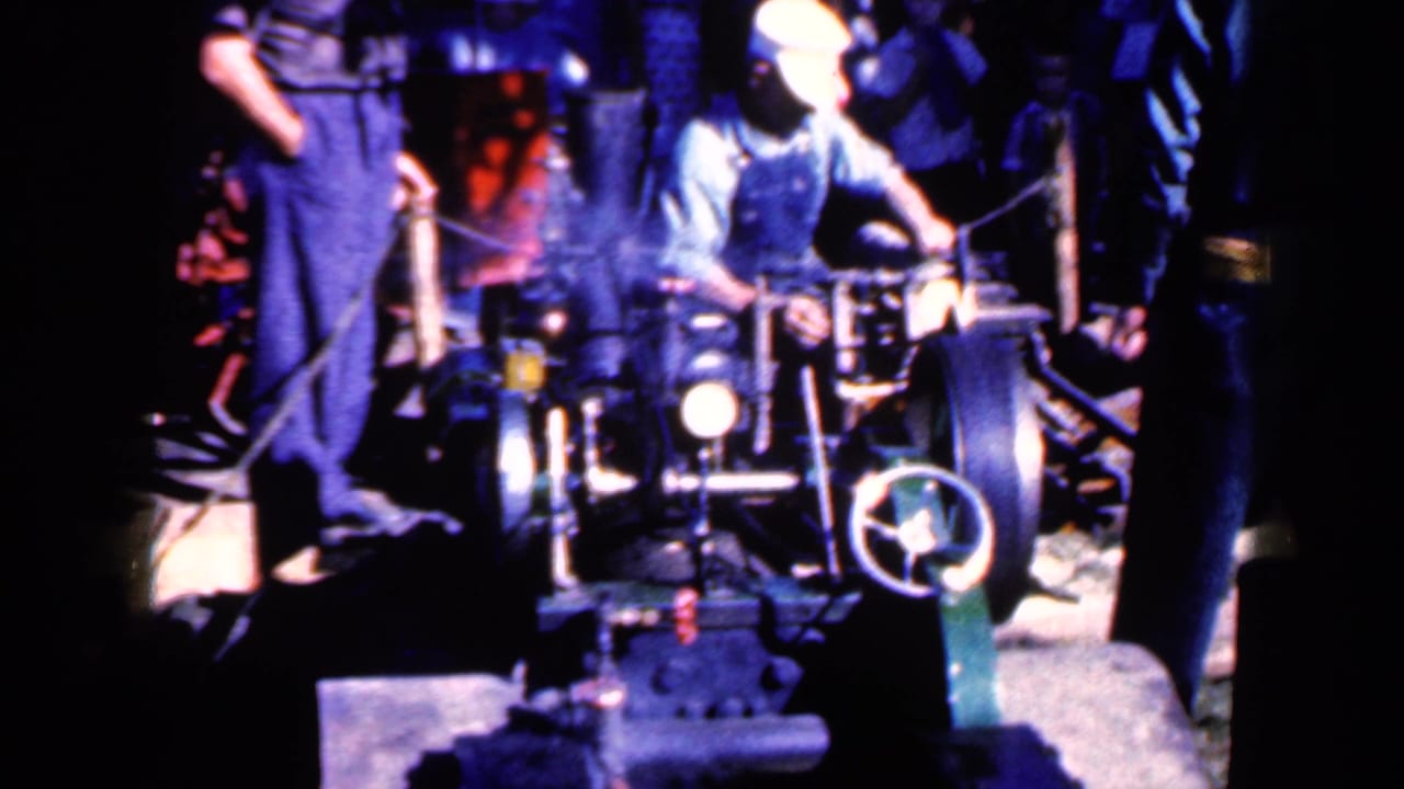 Worker in overalls tends a large factory machine while another worker stands nearby.