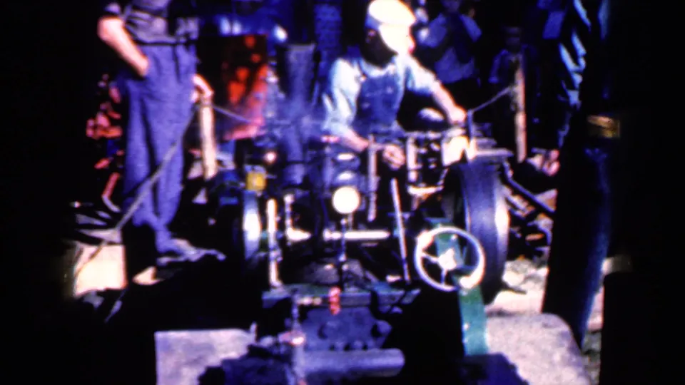 In Iowa 1959 factory footage, a worker in overalls and a white cap operates machinery as another worker stands beside him.