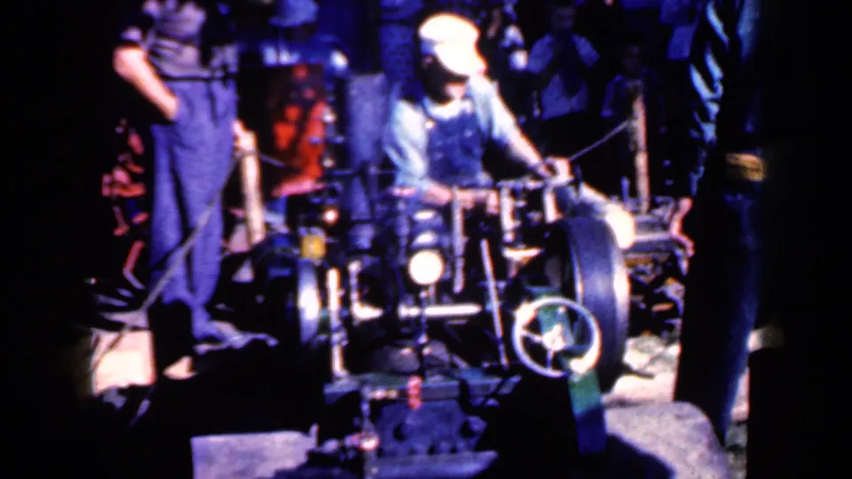 A worker in overalls and cap works beside a prominent handwheel and dark machinery in the supplied 1959 Iowa factory footage.