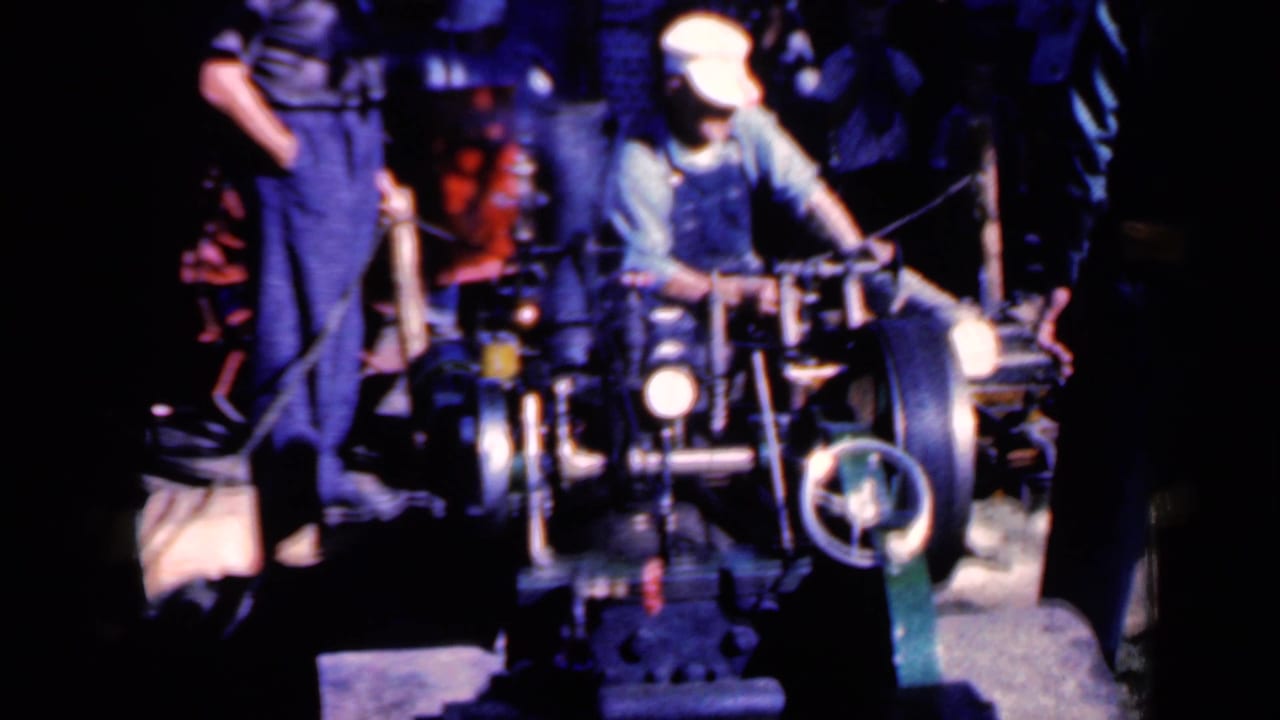 Man in white cap operates a large wheeled machine while another man stands nearby.