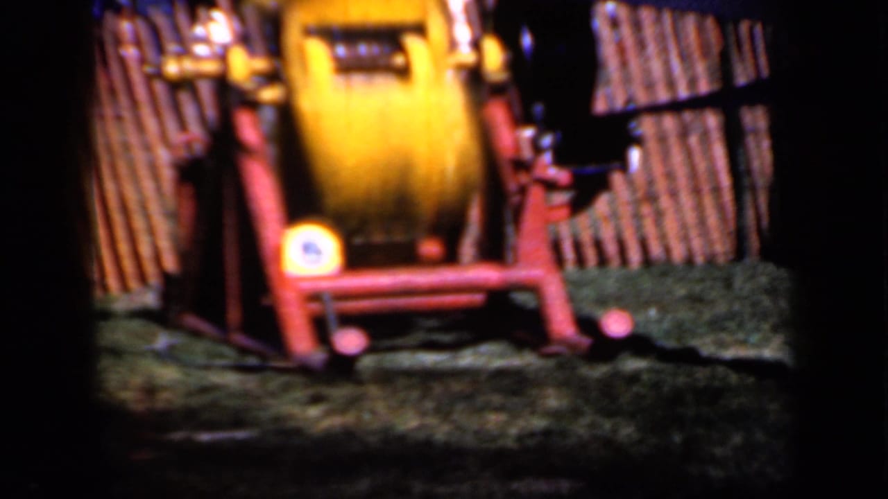 Yellow machine on a red frame operating beside a wooden fence on grass.