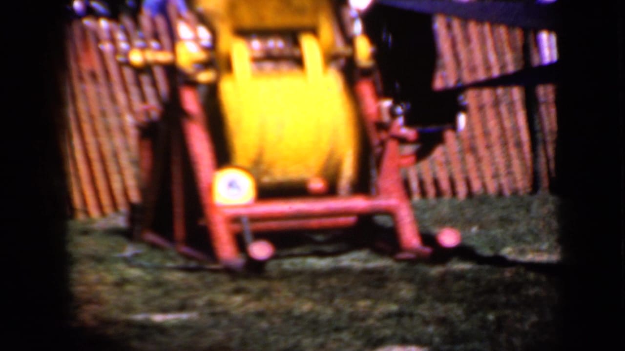 Soft 8mm view of the yellow machine operating in an outdoor fenced area in Iowa, with red supports and a few indistinct people in back.