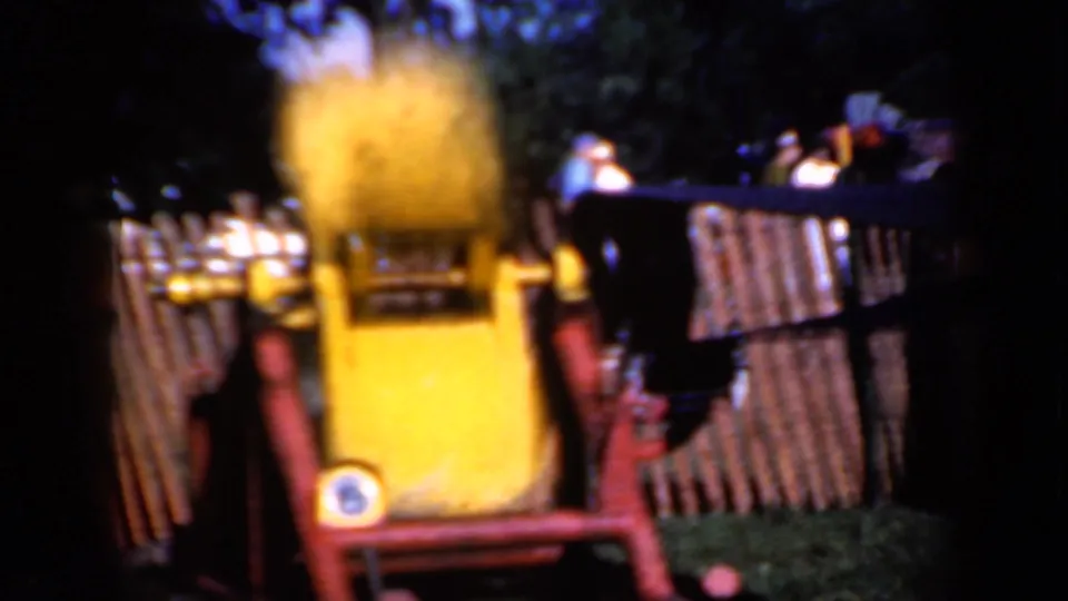 Wider 8mm frame from Iowa in 1959 showing the yellow machine within red supports, wooden fencing, grass, and blurred spectators beyond.
