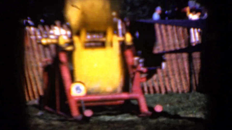 In 1959 Iowa home-movie footage, a yellow rotating machine sits on a red frame near a wooden slat fence.