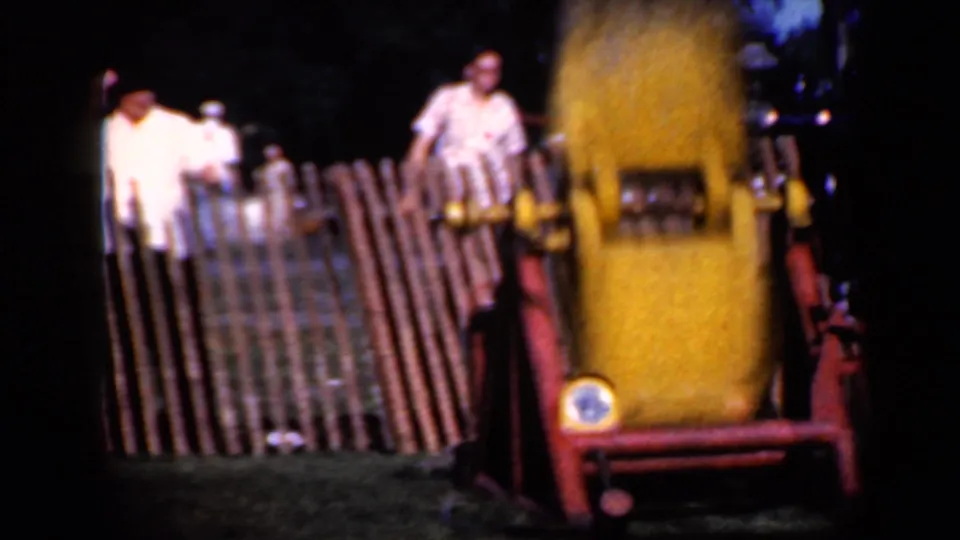 1959 Iowa home-movie frame showing a yellow train-themed attraction near a wooden fence as spectators watch from behind it.