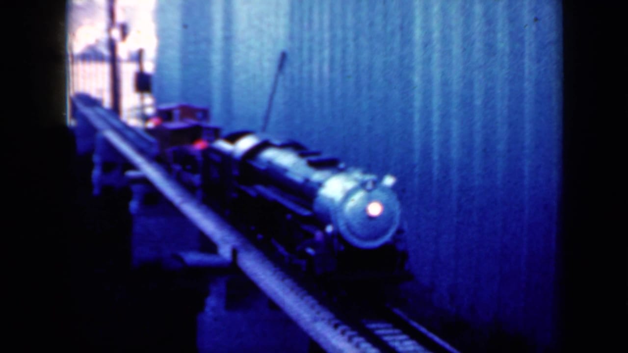 Small steam-style model locomotive approaching on miniature tracks in blue-tinted indoor light.