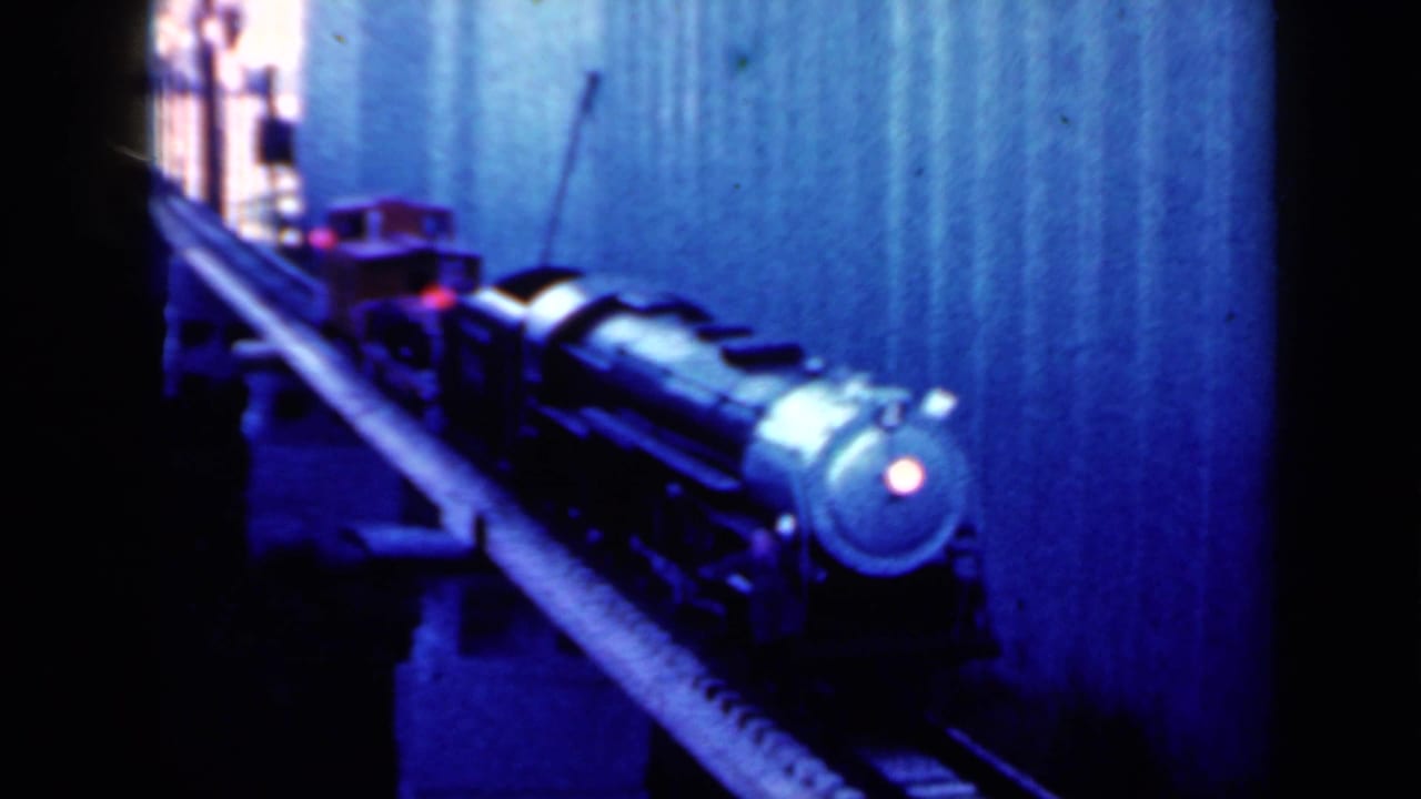 Model locomotive moving toward the camera beside a corrugated wall with a red headlight visible.