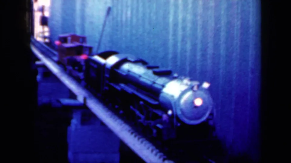 Blue-tinted 1959 Iowa 8mm frame with a small model steam-style locomotive and trailing car on indoor tracks.