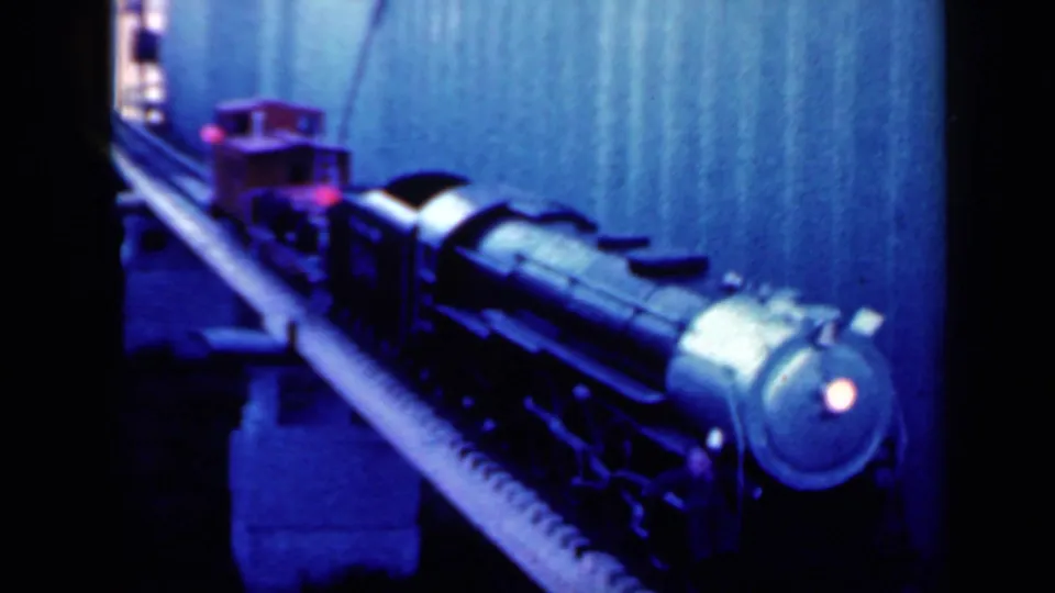 1959 Iowa home-movie frame showing the model locomotive near the camera, blue-tinted with a glowing red front light.