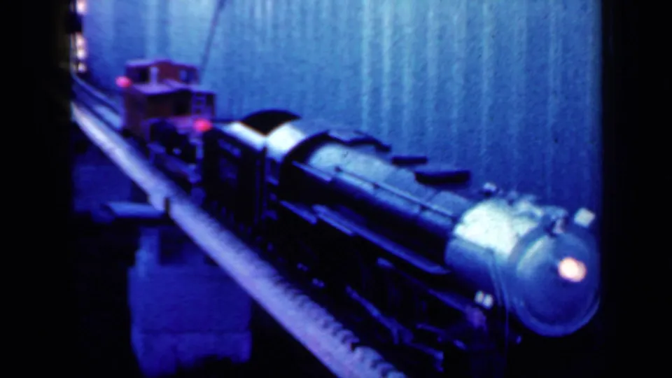1959 Iowa 8mm frame showing a red and black toy train passing along a raised model track in blue indoor light.