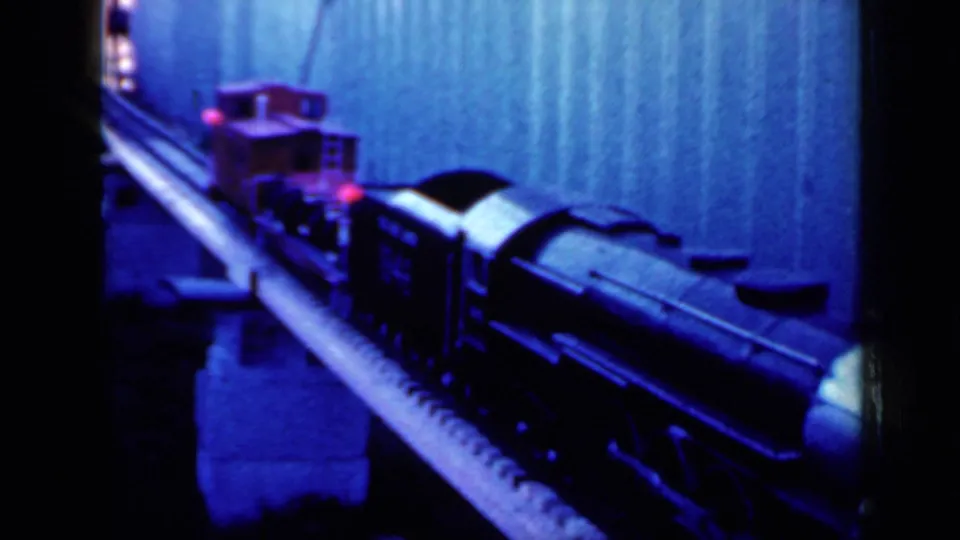 1959 Iowa home-movie frame with the toy train closer to camera, showing dark rail cars and a red car behind them.