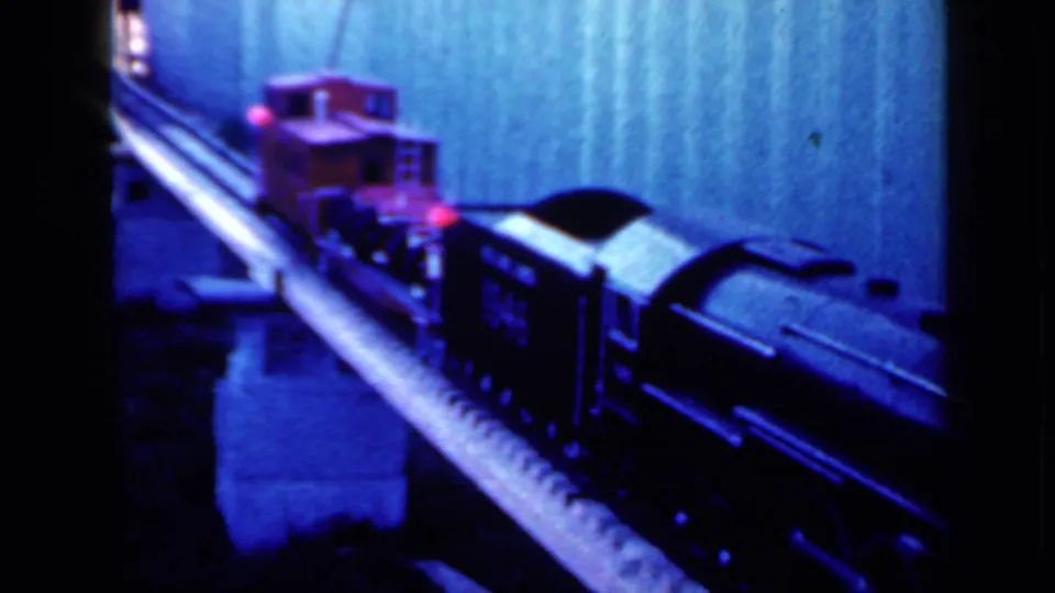 1959 Iowa 8mm frame showing the toy train mid-pass, with the red car and dark locomotive body prominent on the track.