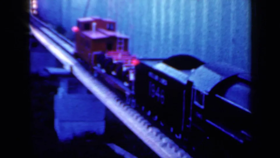 1959 Iowa home-movie frame showing the toy train continuing along the raised track, with red and black cars visible.