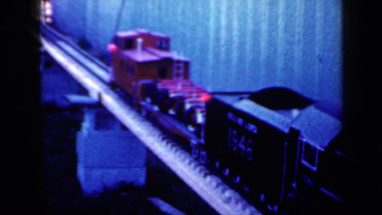 Red model caboose and dark freight cars crossing a narrow miniature bridge.