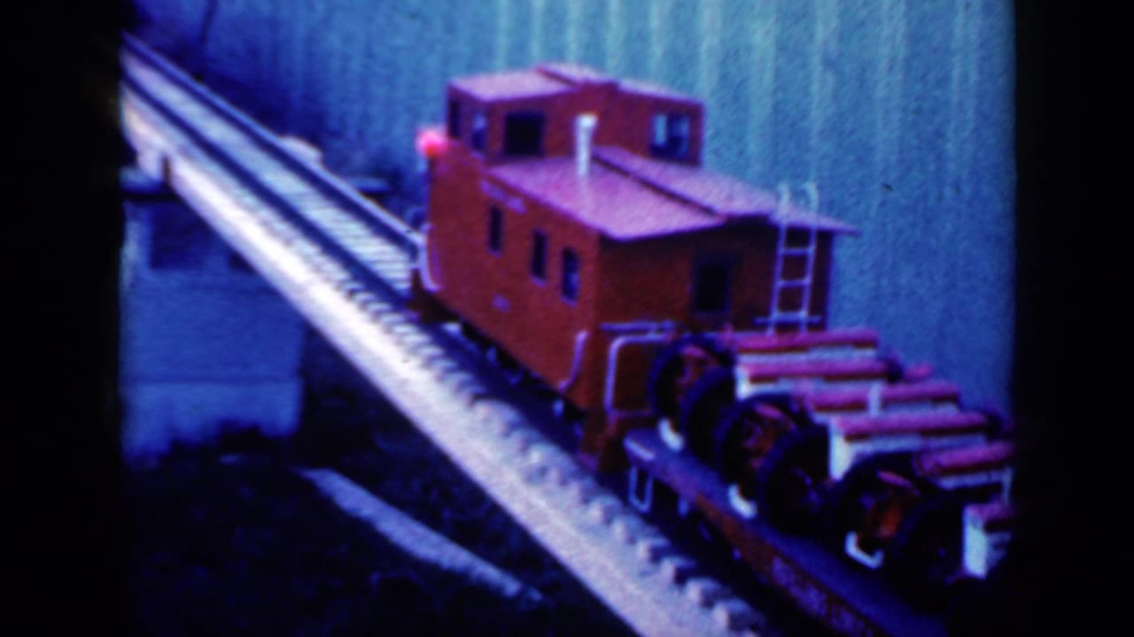 Red model caboose moving along a raised miniature track beside a wall.