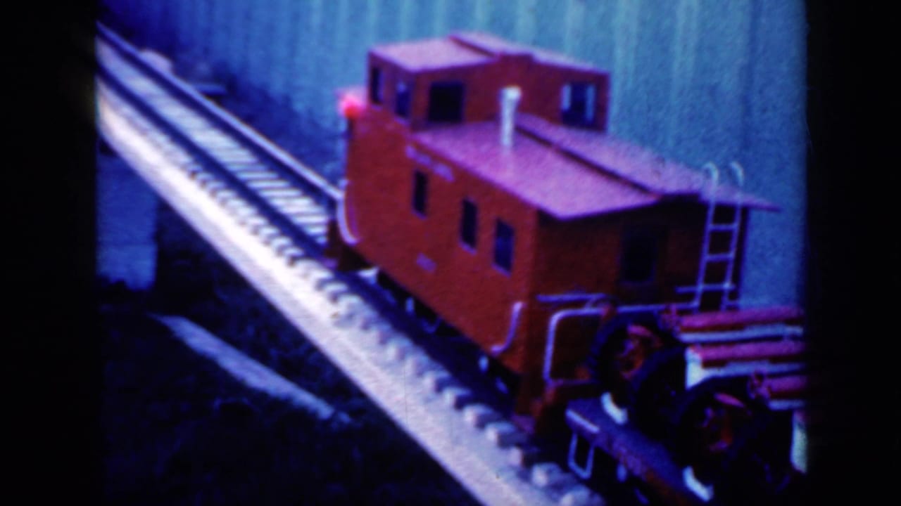 1959 Iowa frame of a red model caboose and cars passing slowly on a raised miniature railway, filmed in saturated 8mm color.