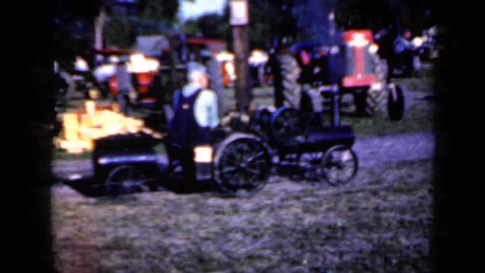 1959 Iowa home-movie frame showing an adult man in overalls near a small steam tractor, with larger tractors behind him at an outdoor exhibition.