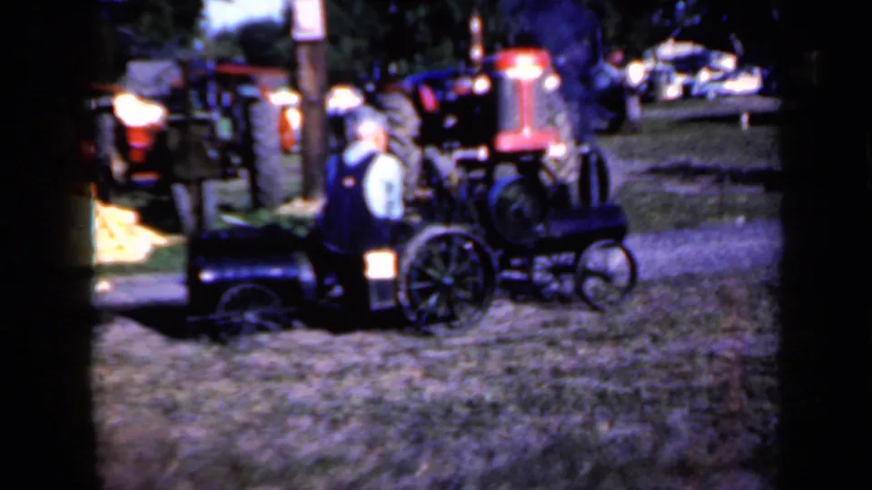1959 Iowa frame of an adult man beside a compact steam-powered tractor, with larger tractors parked behind in a fair-like outdoor setting.
