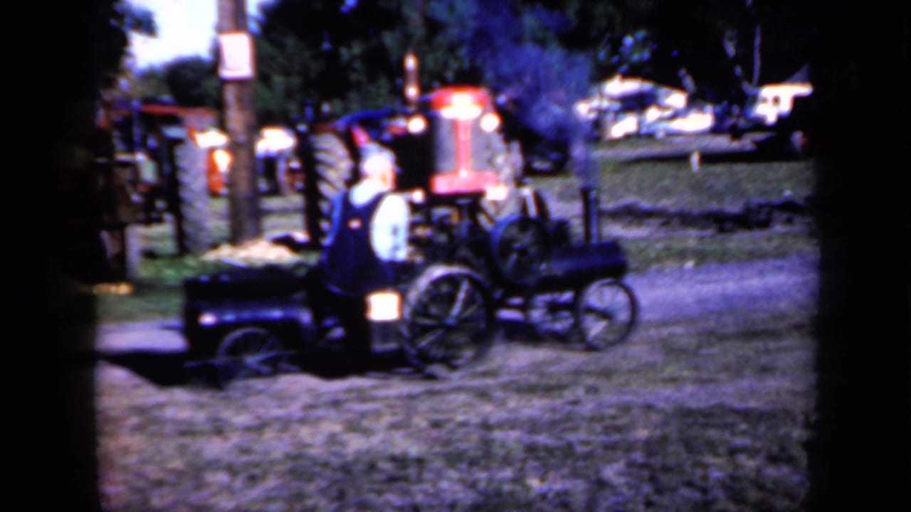 Small steam tractor pulling a cart on grass with an operator and other tractors behind it.