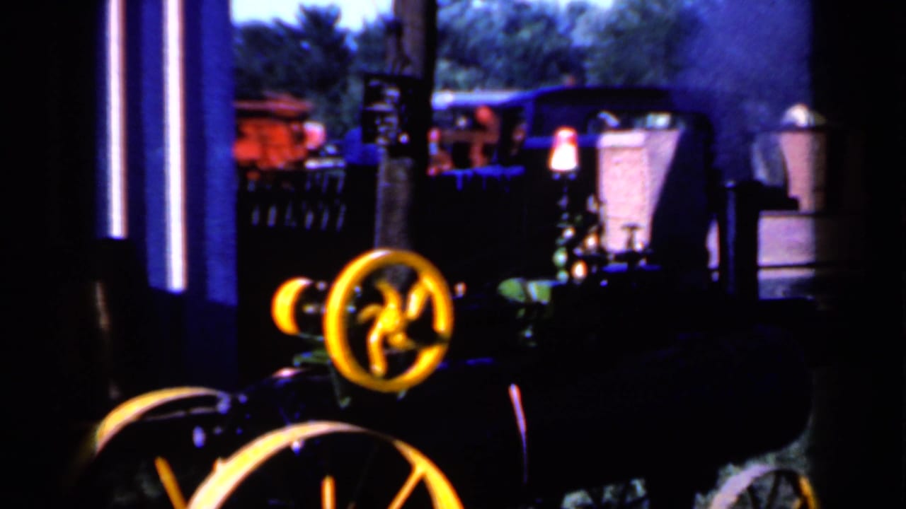 Close 1959 Iowa fairground view of a small steam tractor with a yellow flywheel, dark boiler, and smoke in the background.