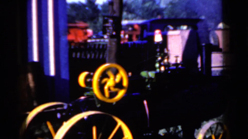 Close home-movie view of the small steam tractor at an Iowa rural fair, with yellow wheels, smoke, and other equipment behind it.
