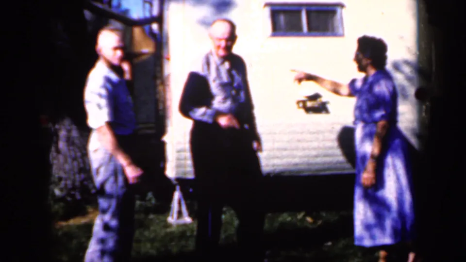 In Iowa in 1959, two men and a woman in a blue patterned dress stand outside a white trailer, apparently talking casually.