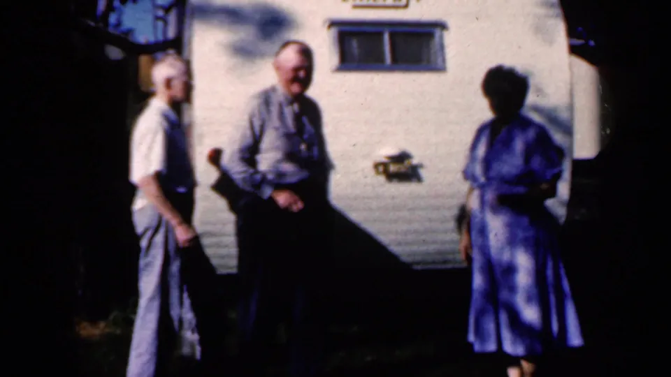 The 1959 Iowa home-movie frame shows three adults standing in conversation beside a white travel trailer with a small window.