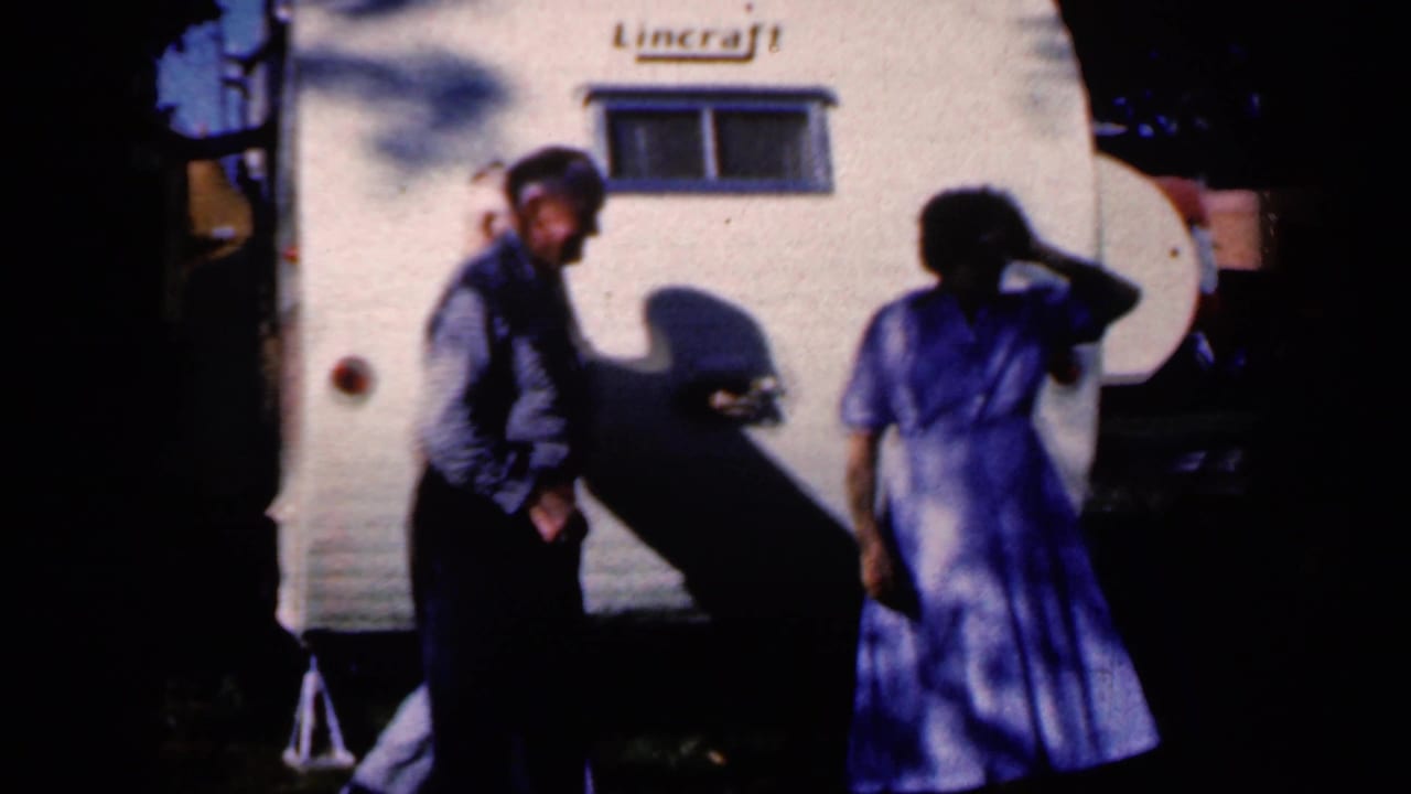 Two adults stand beside a white Lincraft trailer in strong sunlight and shadow.