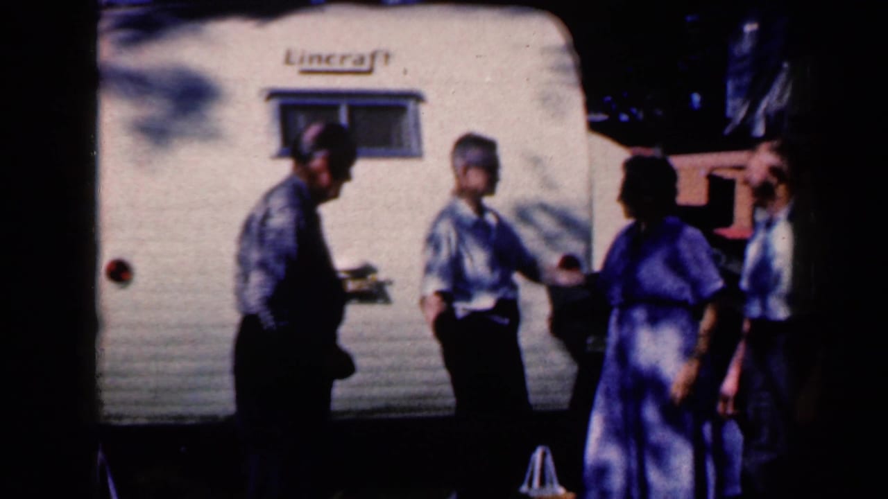 Adults in mid-century clothing stand and talk beside a white Lincraft trailer in the 1959 Iowa clip, with the brand name visible above the window.